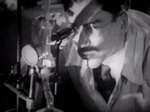 Black-and-white film still of a man studying something through a microscope